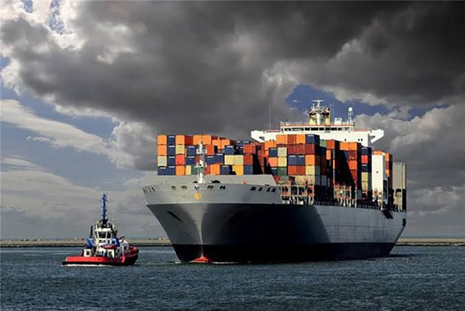 A photograph of a large container ship loaded with colorful shipping containers being escorted by a small red and blue tugboat in open water under cloudy skies.
