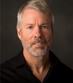 A close-up portrait photo of an older man with grey hair and beard, wearing a dark shirt against a dark background.
