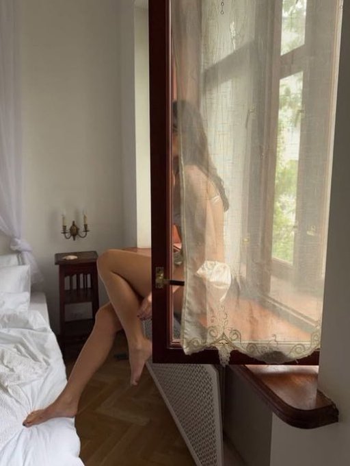 An interior scene showing a woman in a white lace garment sitting by an open window with sheer curtains, with a bed and a bedside table holding candlesticks visible in the softly lit room.
