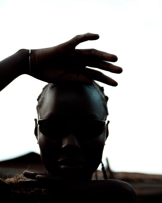 A dramatic silhouetted portrait of a person wearing sunglasses with their hand raised above their head, backlit by bright white light.