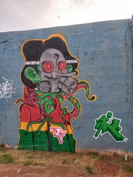 A colorful graffiti mural painted on a blue wall depicting a skull-faced character with red eyes and green tentacles, accompanied by a green stick figure tag.