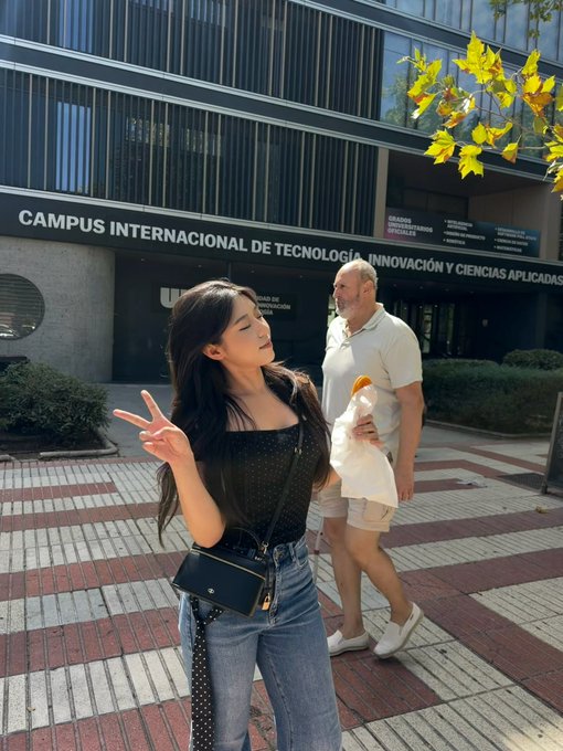 A young woman making a peace sign in front of a building labeled "CAMPUS INTERNACIONAL DE TECNOLOGÍA, INNOVACIÓN Y CIENCIAS APLICADAS", with a man walking behind her.