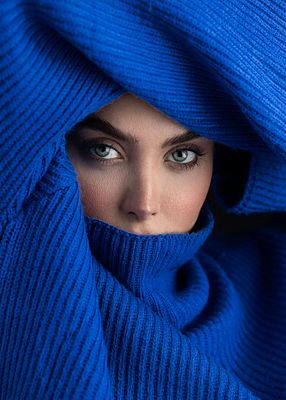 A close-up portrait of a woman with striking blue eyes, her face framed by a vibrant, textured blue knit hood and scarf.