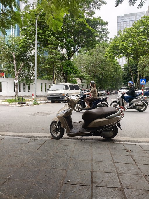 A street scene with a beige motor scooter parked on the sidewalk in the foreground, while two riders on scooters pass by on the road behind it, surrounded by trees and buildings.