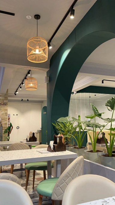 An interior view of a restaurant or café featuring dark green arched walls, hanging woven pendant lights, white marble tables, and white chairs with green cushions, decorated with potted plants.