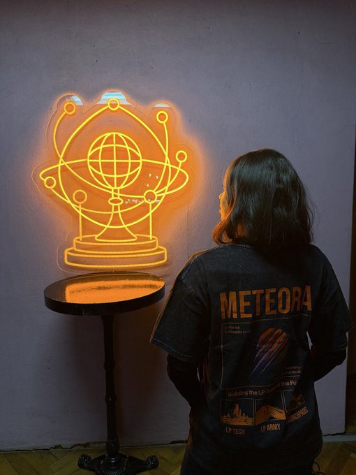 Person wearing a 'METEORA' graphic t-shirt standing before an orange neon sign depicting a crystal ball with an atomic-style globe inside it.