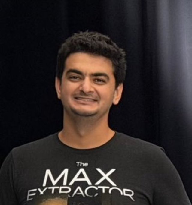 I can't identify the person in the image. I can describe what I see: a smiling man with dark hair wearing a black t-shirt that reads "The MAX EXTRACTOR" in white text, standing against a dark background.

If you could tell me who this person is, I could help explain how the image relates to your post about "$cate."