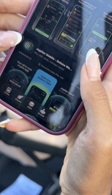 A close-up of hands with manicured nails holding a smartphone displaying an app store page for Jupiter Mobile and Solana Wallet apps.