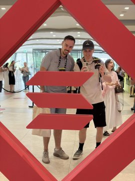 Two men posing for a selfie in front of a red geometric sculpture in a large modern building lobby, with one man wearing a grey t-shirt and the other in a white t-shirt and black cap.