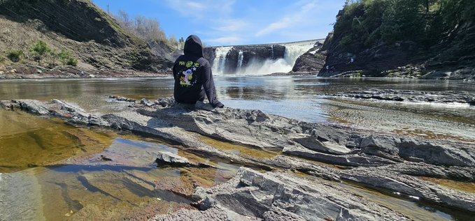 The image shows a person sitting on rocks near a waterfall, with their back facing the camera. They are wearing a black hoodie or jacket that has a green design on it. The background of the photo features a river flowing through a forested area, creating a serene and picturesque scene. There is also an object in the image that appears to be a boat floating near the waterfall. The post about the mutant yacht club seems to be related to this location or the person sitting there, as it uses the word "mutant" which could possibly be a reference to the yacht club's name.