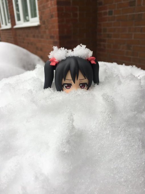 An anime figure with black twin tails and pink ribbons is partially buried in a pile of snow, with only its head visible against a brick wall background.