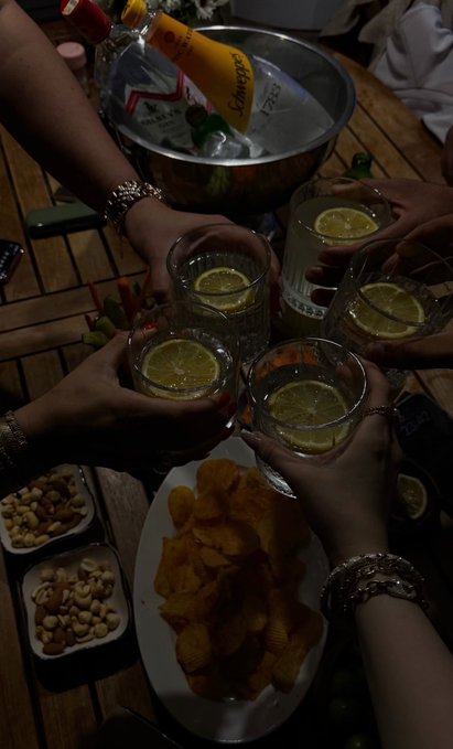 A group of people toast with glasses of lemon-infused drinks around a wooden table with snacks, a champagne bottle, and a bowl of ice.