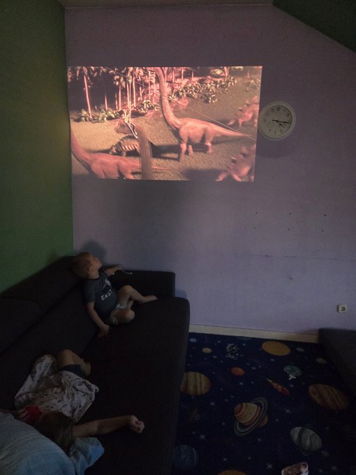 Two children relax on a dark couch while a projected image of dinosaurs from Jurassic Park plays on the lavender wall above them, with a wall clock showing approximately 9:30 nearby. The cozy, dimly lit room features a space-themed rug depicting planets, creating a perfect pre-event atmosphere for the JurassicFi fundraiser warmup with the kids.