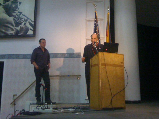 Two men on a stage, with one speaking at a wooden podium and the other standing nearby, next to a framed black-and-white portrait.