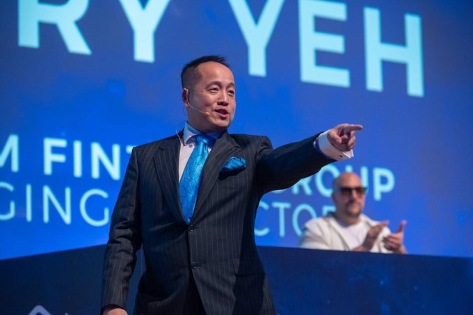 A photograph of a man in a dark pinstripe suit with a blue tie and pocket square speaking on stage, gesturing with his arm, with partial text "ARY YEH" visible on a blue screen behind him.