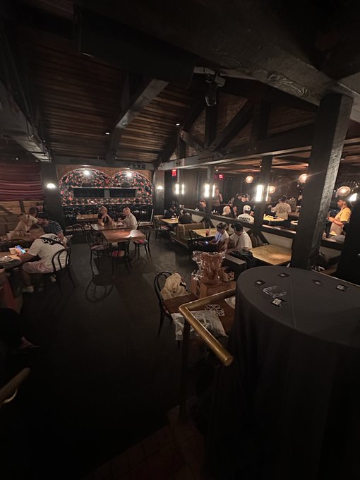 Interior view of a dimly lit bar or lounge with exposed wooden ceiling beams, patterned tile walls, and patrons seated at tables and the bar.