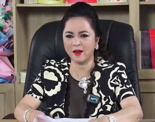 A photograph of a Vietnamese businesswoman wearing a black and white patterned blazer with a diamond brooch, seated in a black office chair reading documents.