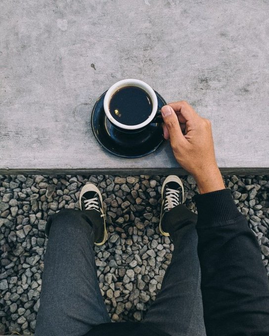 In this image, a person wearing black pants and sneakers is sitting on a concrete ledge or step, holding a cup of coffee. The background features a gray stone wall, which adds an urban feel to the scene. There are also some rocks visible in the area where the person is seated. The photo appears to be taken from above, providing a unique perspective that highlights the person and their surroundings.