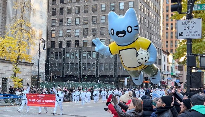 A parade scene on a city street showing a large inflatable balloon shaped like a blue cat character in yellow clothing, with people walking below holding a banner that reads 'BLUE CAT & CHUBS' while spectators watch.