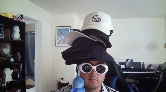 A man sits indoors wearing a stack of multiple hats, large white-rimmed sunglasses, and holding a small blue plush toy.