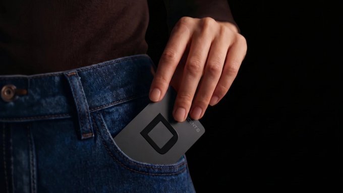 The image shows a person's hand placing a sleek black card-style wallet from D'CENT into the back pocket of their blue jeans, with the brand's diamond logo visible on the card. The partial text "WAL" can be seen on the device, highlighting its slim, credit-card-sized design that fits easily into a pocket for everyday carry.