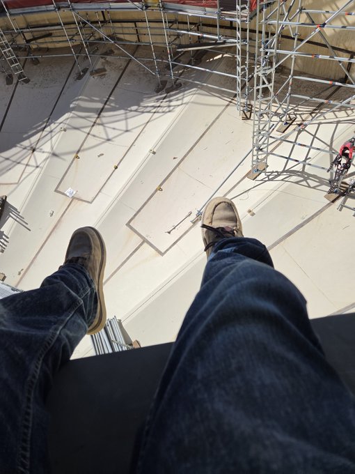 First-person perspective photo looking down at legs wearing jeans and tan work boots dangling from a high elevation above a construction site with scaffolding and large white panels below.