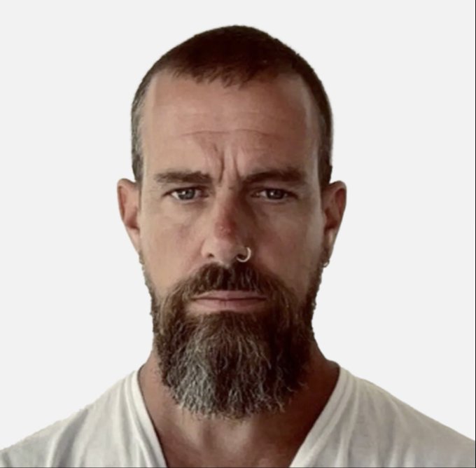 A portrait photograph of a serious man with a graying beard, short hair, a nose ring, and ear piercings, wearing a white t-shirt against a white background.
