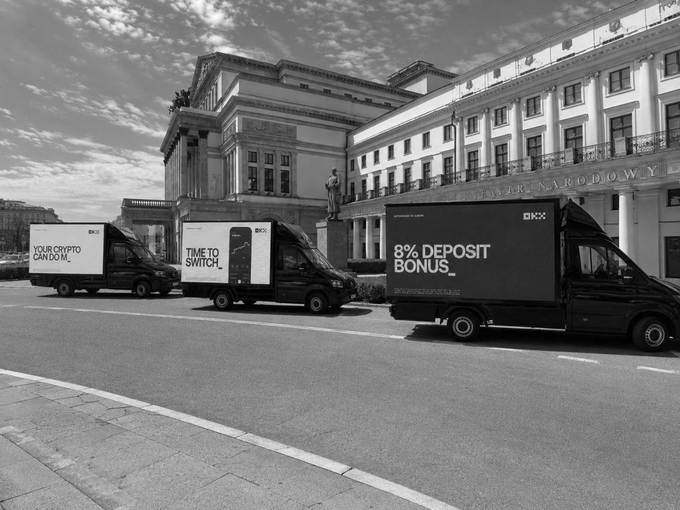 A black-and-white photo of three OKX delivery vans with messages like "YOUR CRYPTO CAN DO M..." and "8% DEPOSIT BONUS" parked in front of a classical building in Warsaw.