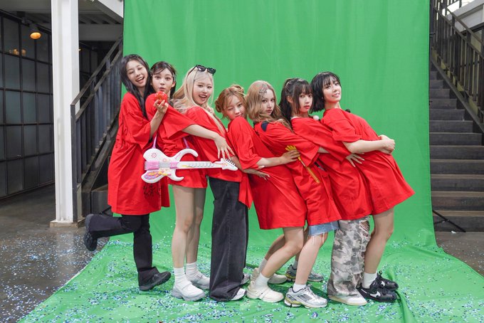 A group of eight people wearing matching red outfits pose together on a green screen set, with one holding a pink guitar prop, surrounded by scattered confetti.