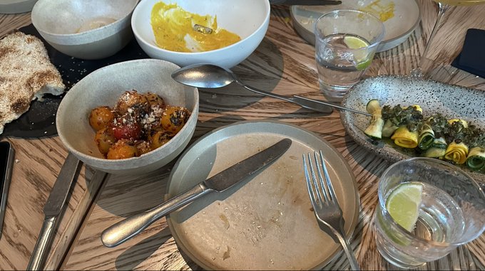 A dinner table setting featuring a spread of dishes including bread, roasted vegetables, and a green salad with cucumber and avocado, accompanied by water glasses with lime. The meal reflects the post's context of a networking dinner with OKX EU and Product teams during the Dutch Blockchain Week.