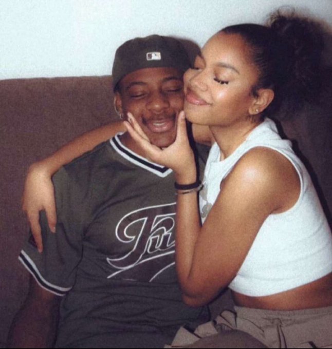 A young couple shares a tender moment on a couch, with the woman in a white tank top affectionately cradling the man's face as he wears a gray "JR" jersey and a baseball cap. The warm, intimate scene fits the post's casual, playful mid-week vibe of enjoying a productive day with loved ones.