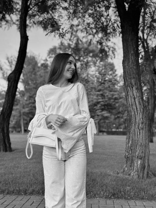 A black-and-white photo of a smiling woman in a light blouse holding a small handbag in a park setting.