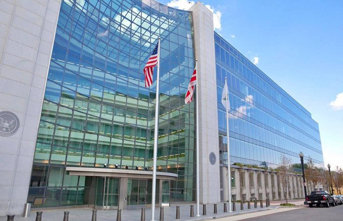 An exterior shot of a modern, multi-story glass building, likely a U.S. government facility, with an American flag and another flag flying on flagpoles in front.