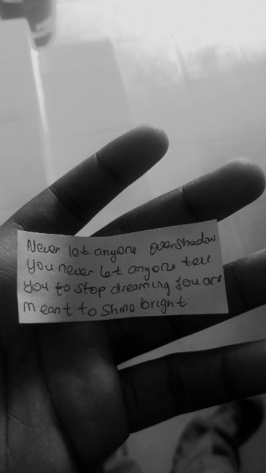 A black and white photo of a hand holding a handwritten note that reads 'Never let anyone Dear Shadow You never let anyone tell you to stop dreaming you are meant to shine bright'.