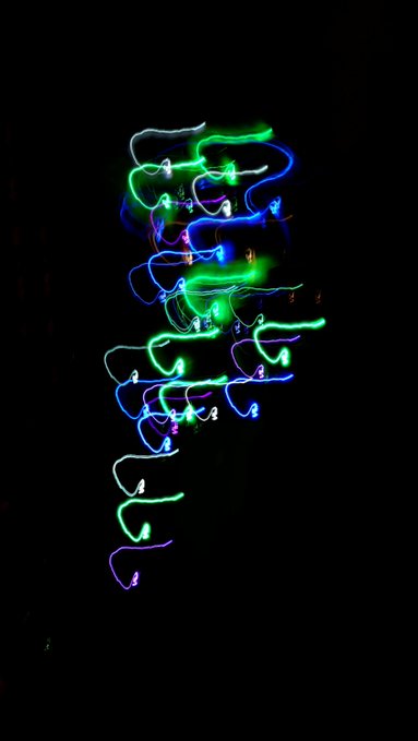 A long-exposure photograph capturing swirling streaks of blue, green, and purple light painting against a pitch-black background.
