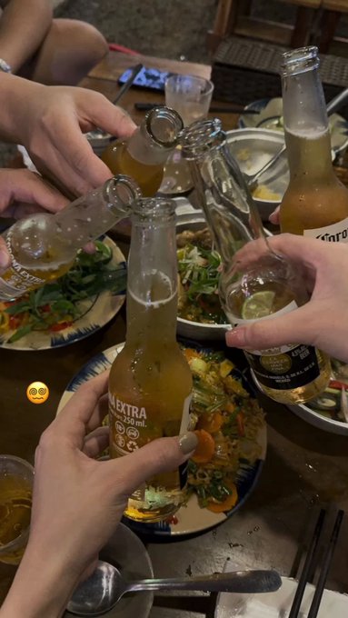 A group of friends clinks bottles of "NEXTRA 330 mL" beer over plates of shared Vietnamese food, celebrating a casual night out. The scene of friends gathered around a lively dinner table contrasts with the post's focus on a quiet, solitary evening spent researching post-quantum security with @quipnetwork.