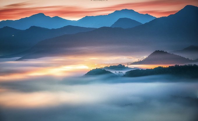 A serene landscape photograph showing misty mountains at sunset with orange and pink hues reflecting off fog-covered valleys.