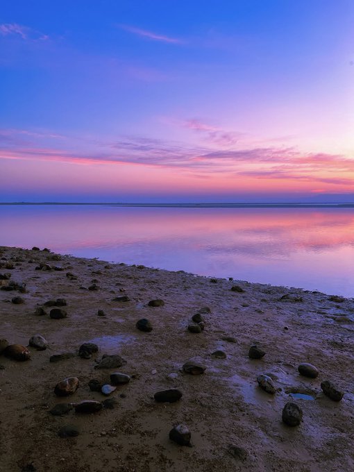 A serene beach scene at sunset or sunrise, featuring a rocky, wet shoreline in the foreground and a calm body of water reflecting pink, purple, and blue hues from the colorful sky.