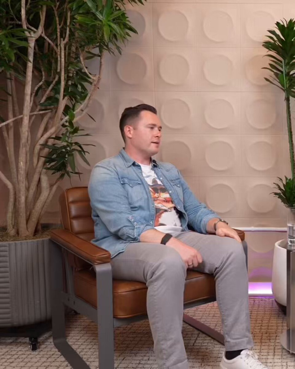 The image shows a man sitting in a chair with his legs crossed, wearing a denim shirt and gray pants. He has a potted plant behind him and another one near a white pillar. The background features a wall adorned with circular patterns that resemble circles of light. There is also a computer monitor on the right side of the room.