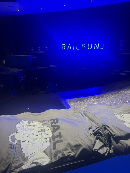 The image shows a neon sign for "Railgun", which appears to be in a dark room with a blue background. The sign is lit up with blue neon light that stands out against the darkness of the room. A person is sitting on top of a mattress placed on a black bed frame, and there are several T-shirts laid out next to them. One of these shirts has a picture of a cat on it, adding a playful touch to the scene. The overall atmosphere seems mysterious as if someone left the room with an item, like a T-shirt or a cigar, for you to discover in this dimly lit environment.