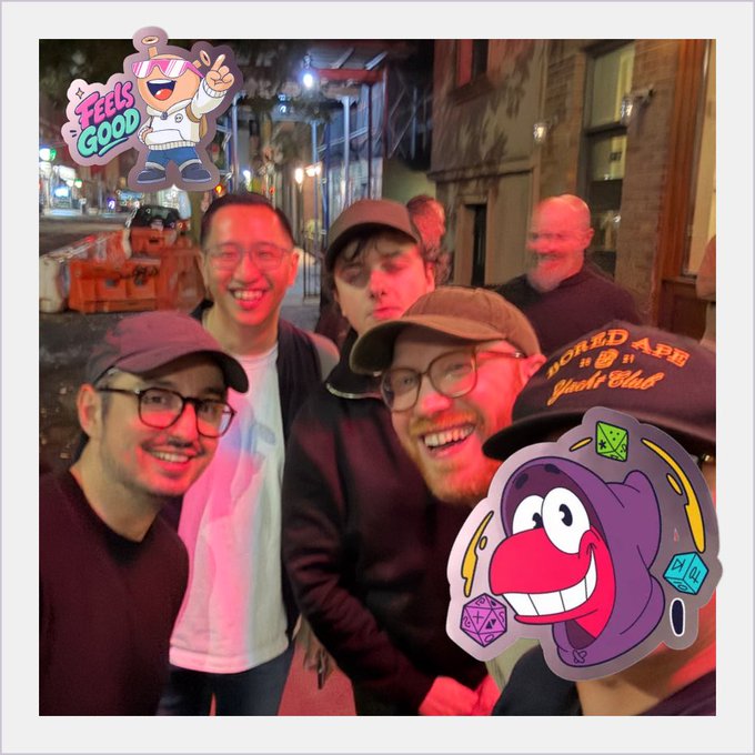 A group of five men taking a selfie on a nighttime city street, decorated with cartoon stickers of a character saying 'Feels Good' and a purple creature with dice.
