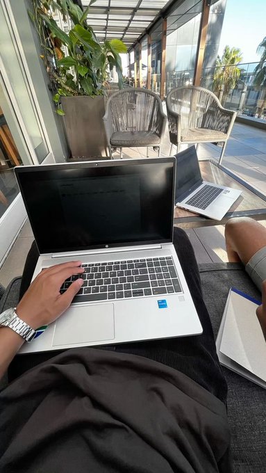 A person works on an HP laptop on their lap in an outdoor lounge area, with another laptop visible on a table nearby.