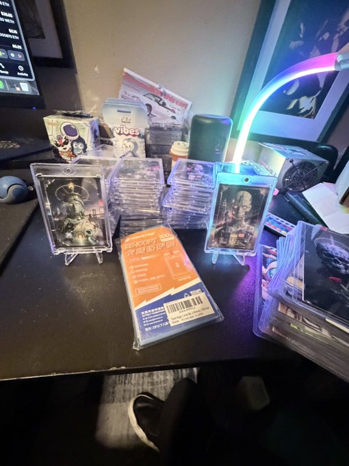 A collection of trading cards and collectibles stacked on a dark desk, including cards in protective cases featuring character artwork, an RGB LED light strip, and various packaged items.