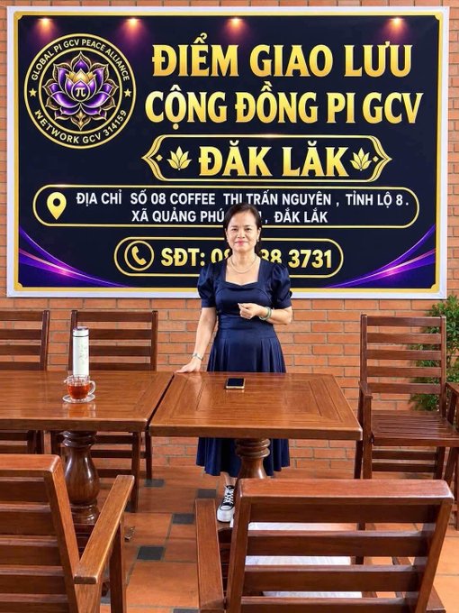 A woman in a dark blue dress stands between wooden tables in front of a banner with Vietnamese text reading 'ĐIỂM GIAO LƯU CỘNG ĐỒNG PI GCV ĐẮK LẮK' and contact details including a phone number and address.