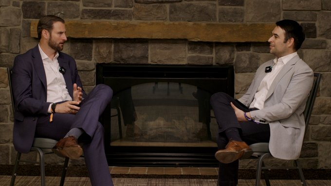 Two men in suits sit facing each other in front of a stone fireplace, appearing to be engaged in a conversation or interview, both wearing lapel microphones.