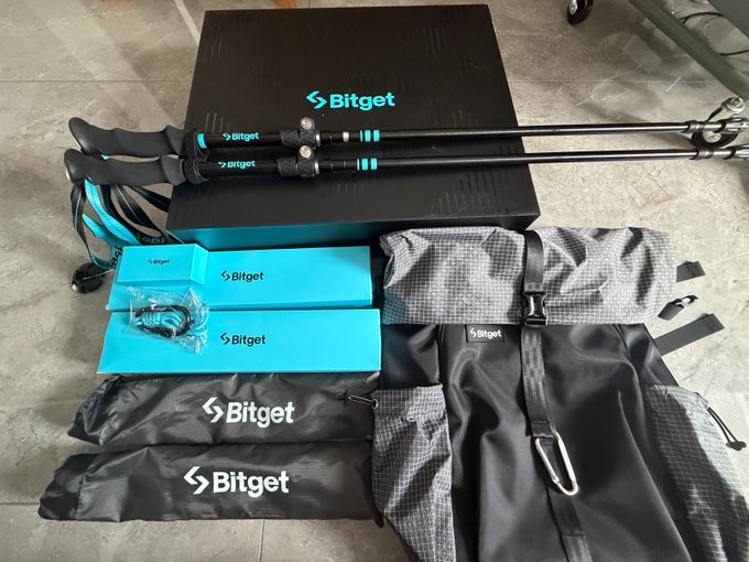 Overhead shot of Bitget-branded merchandise including trekking poles, boxes, a backpack, drawstring bags, and carabiners arranged on a concrete floor.