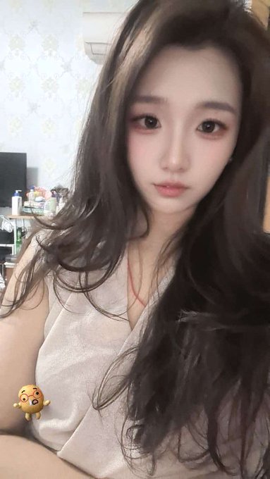 A young woman with long dark hair poses for a close-up selfie in what appears to be a bedroom, using a crying baby emoji sticker to humorously illustrate her relatable post about having a cheerful personality due simply to having a bad memory rather than the privileged reasons others might have.
