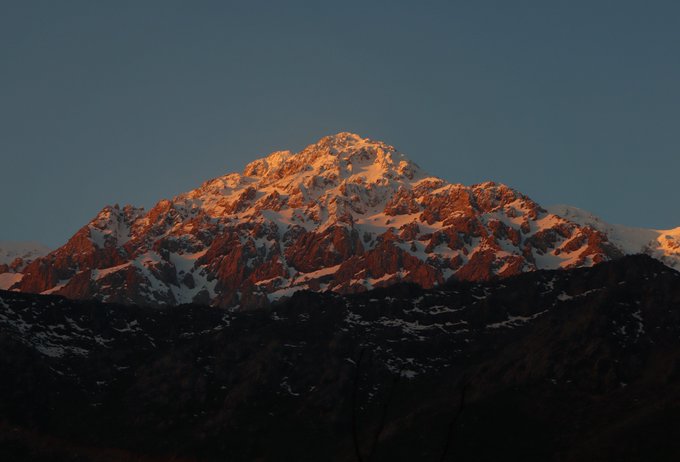 Snow-capped mountain peak glowing orange in alpenglow at sunset or sunrise against a deep blue sky.