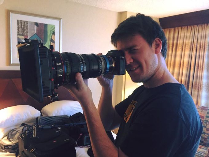 A man smiles while holding a professional cinema camera with a large lens, standing in a hotel room.