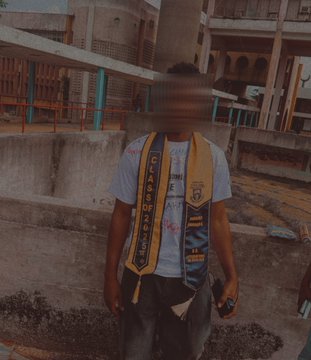 A person wearing a gray t-shirt and a yellow and blue scarf standing outdoors in front of a concrete structure, with their face obscured by a blur effect.